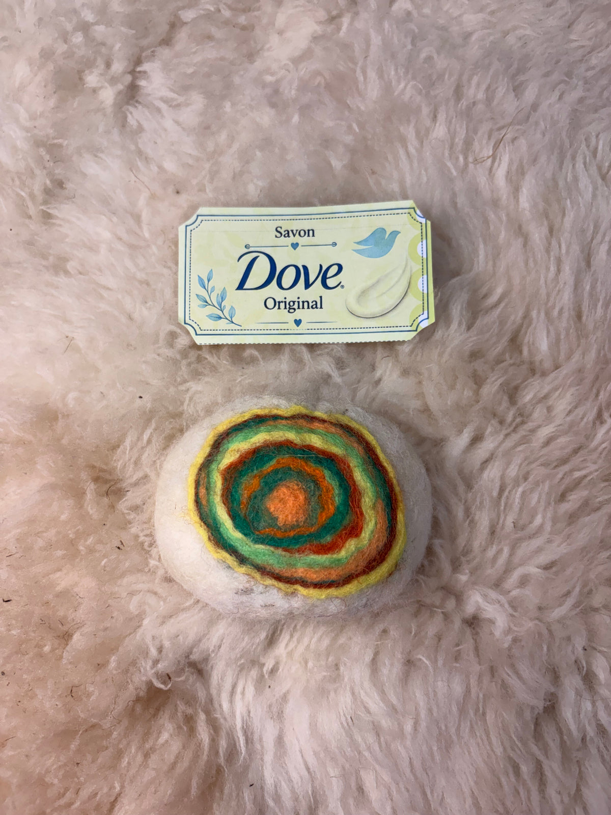Savon feutré artisanal en laine naturelle au motif de cible en spirale orange, verte et jaune, posé sur une toison de mouton crème, étiquette Dove Original au-dessus.